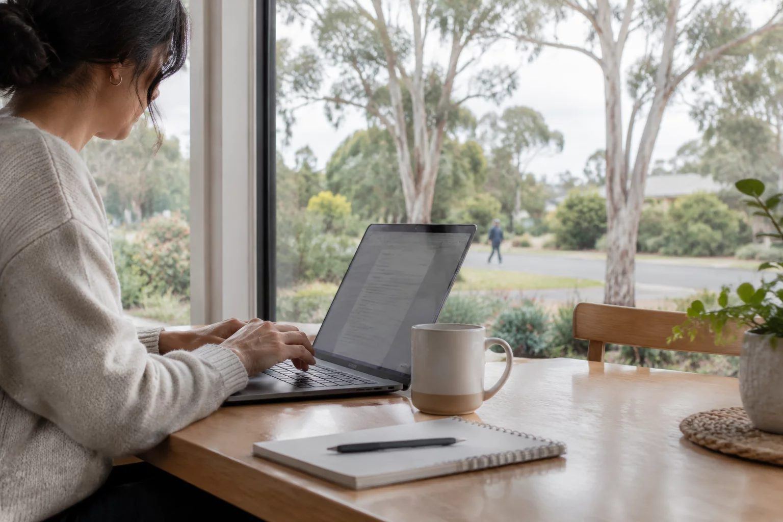 A person working on a laptop at a home desk by a window